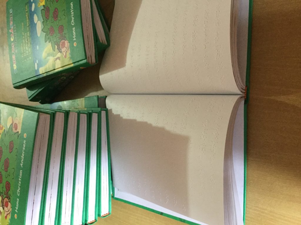 Foto en el que se ve una Pila de libros Braille junto a un libro abierto